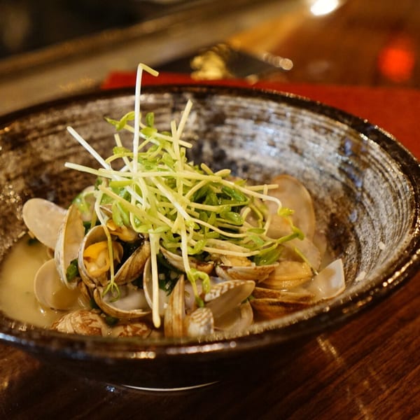 Clams in a dark bowl with broth, topped with fresh green sprouts on a wooden table.