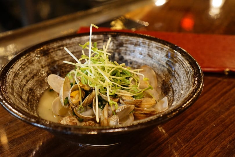 Clams in a dark bowl with broth, topped with fresh green sprouts on a wooden table.