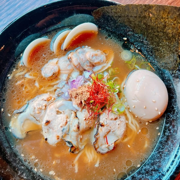 Seafood ramen with oysters, clams, a soft-boiled egg, and seaweed in a black bowl.