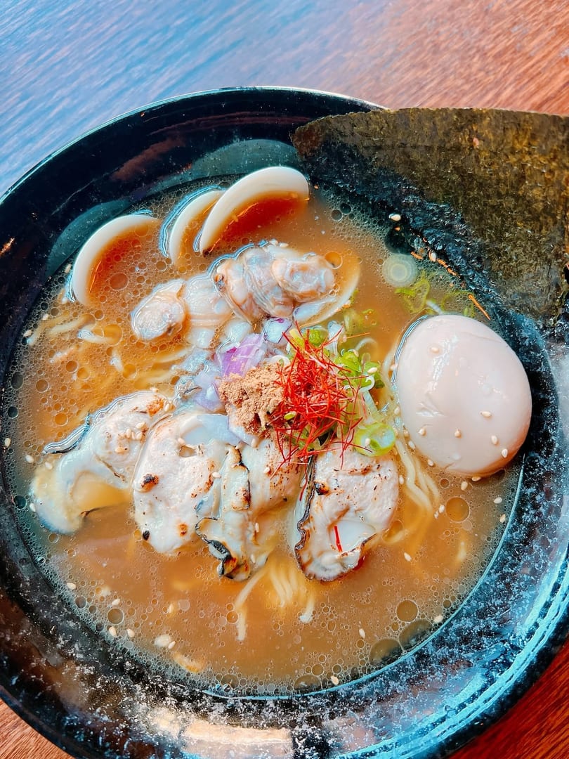 Seafood ramen with oysters, clams, a soft-boiled egg, and seaweed in a black bowl.