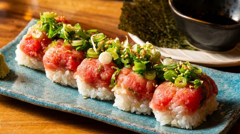Five pieces of nigiri sushi with minced red tuna, green onions, and sesame seeds on a blue plate.