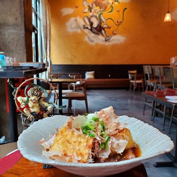 A dish of fried tofu with bonito flakes and scallions, in a restaurant with a mural.