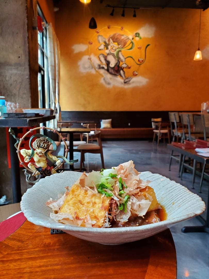 A dish of fried tofu with bonito flakes and scallions, in a restaurant with a mural.