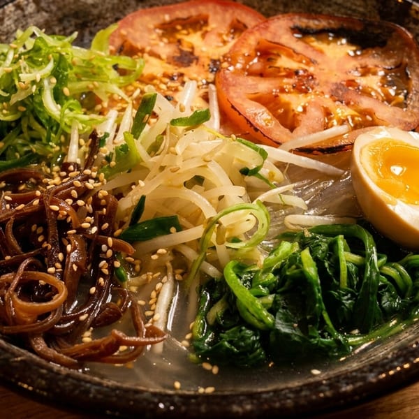 A close-up of a ramen bowl with noodles, grilled tomatoes, bean sprouts, spinach, and a soft-boiled egg.