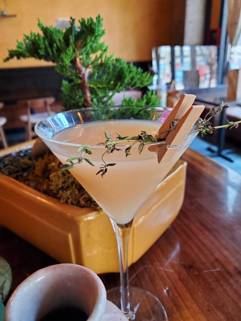 A light-colored martini in a stemmed glass, garnished with herbs and a wooden clip, next to a bonsai.
