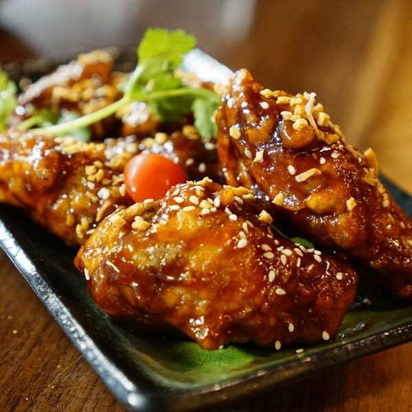 Close-up of glazed chicken wings topped with sesame seeds and peanuts, garnished with cherry tomatoes and cilantro.