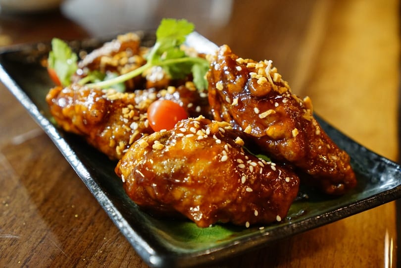 Close-up of glazed chicken wings topped with sesame seeds and peanuts, garnished with cherry tomatoes and cilantro.