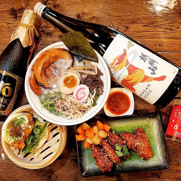 A spread of Japanese dishes including ramen, chicken wings, bao buns, and two bottles of sake.