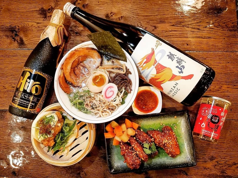 A spread of Japanese dishes including ramen, chicken wings, bao buns, and two bottles of sake.
