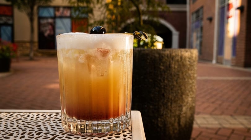 A foamy, layered cocktail with a dark cherry on top, on an outdoor patio table.
