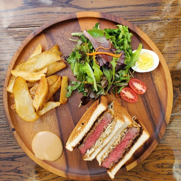 Wagyu katsu sandwich with fries, salad, tomatoes, and a hard-boiled egg on a wooden plate.