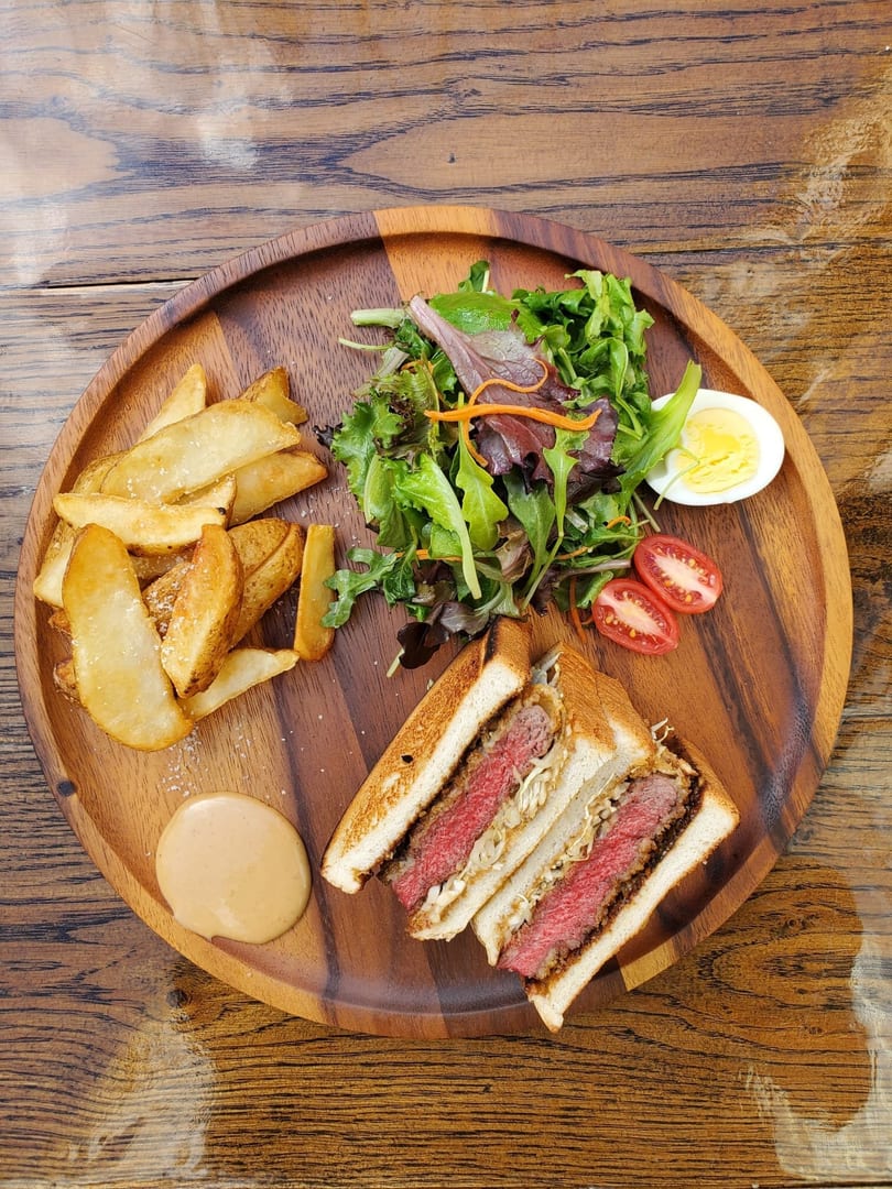 Wagyu katsu sandwich with fries, salad, tomatoes, and a hard-boiled egg on a wooden plate.