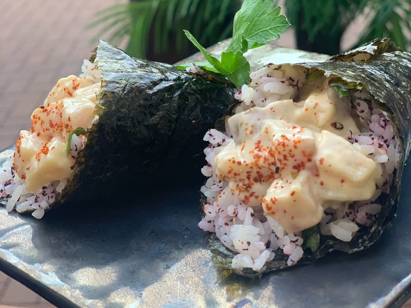 Two temaki hand rolls with creamy filling, rice, and red spice on a dark plate.