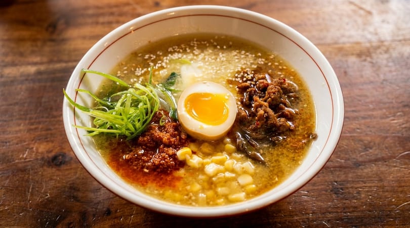 A bowl of ramen with a soft-boiled egg, corn, ground meat, and green onions on a wooden table.