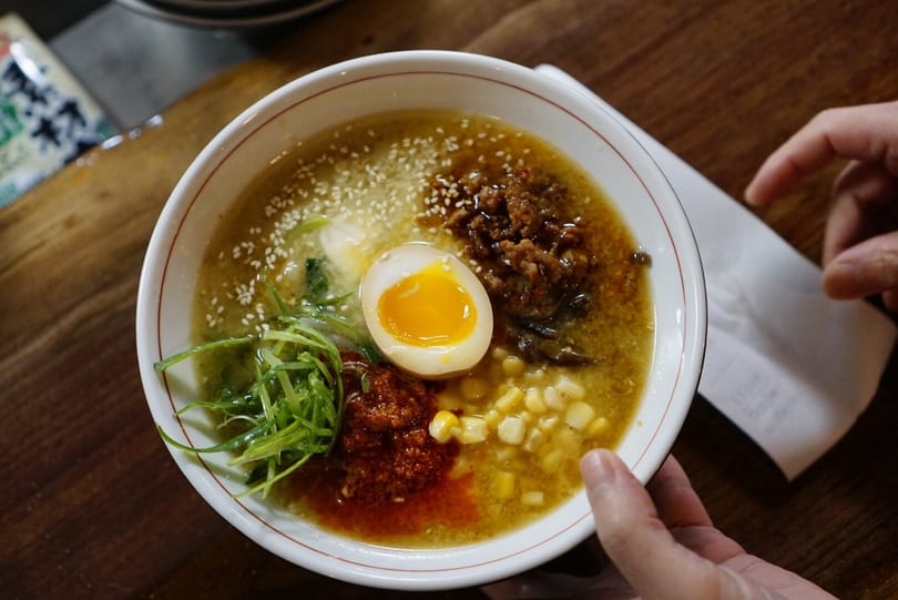 Overhead view of a ramen bowl with a soft-boiled egg, corn, scallions, and ground meat.