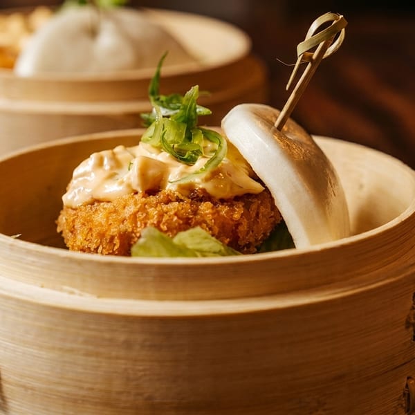 Crispy fried bao bun with sauce and scallions in a bamboo steamer on a wooden table.
