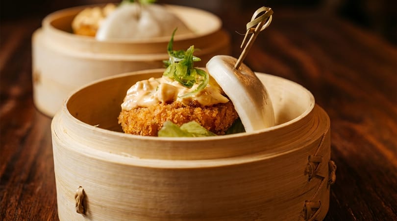 Crispy fried bao bun with sauce and scallions in a bamboo steamer on a wooden table.