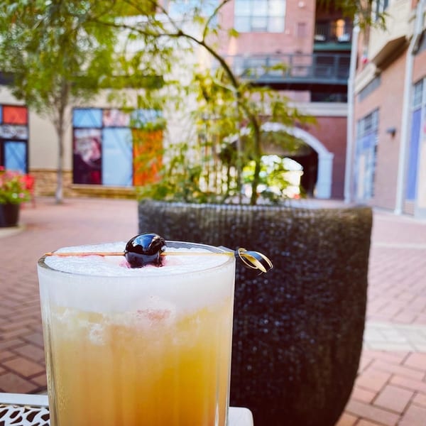 A foamy cocktail with a cherry garnish sits on an outdoor patio table with buildings in the background.