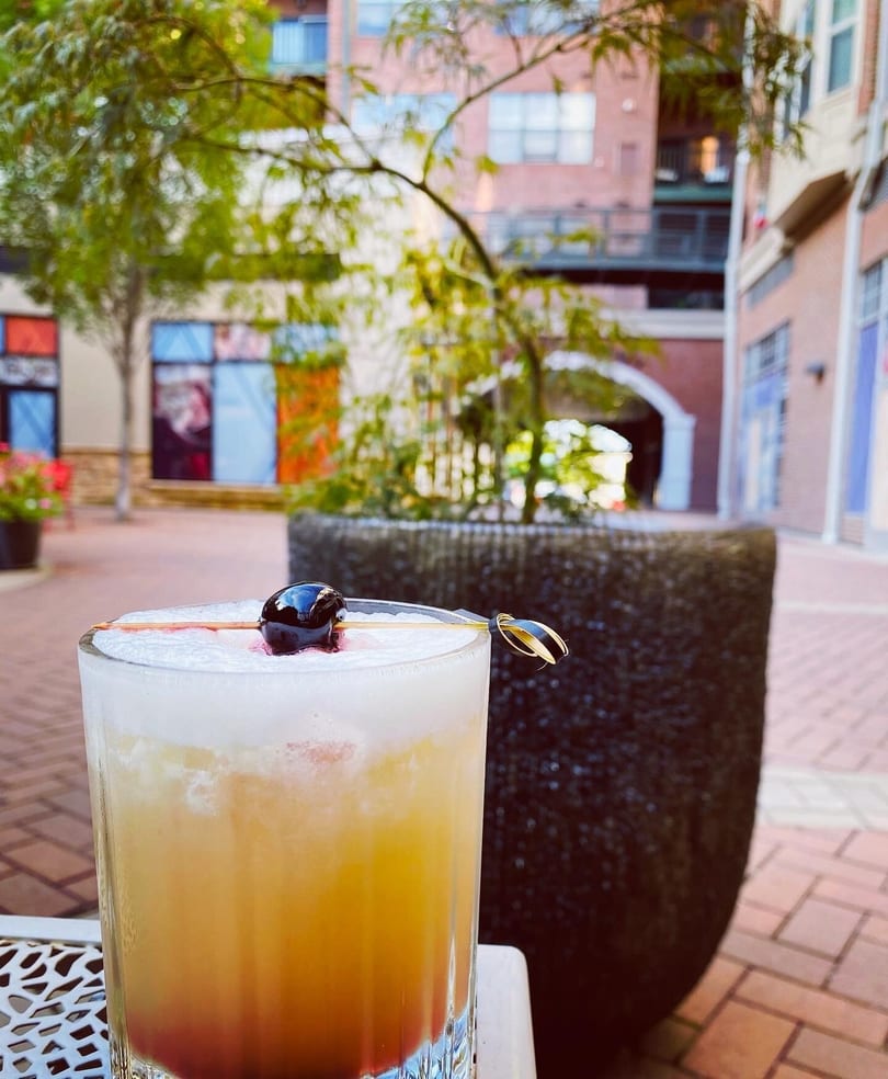 A foamy cocktail with a cherry garnish sits on an outdoor patio table with buildings in the background.